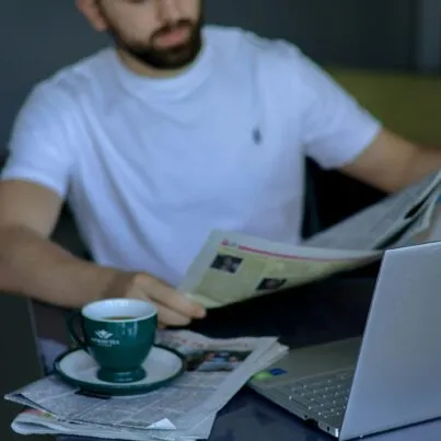 A man reading a newspaper. In front of him is a cup of coffee and a laptop.