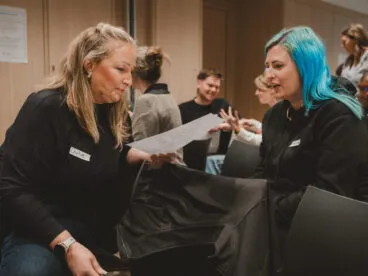 Two people look over a sheet of paper in discussion with others in the background